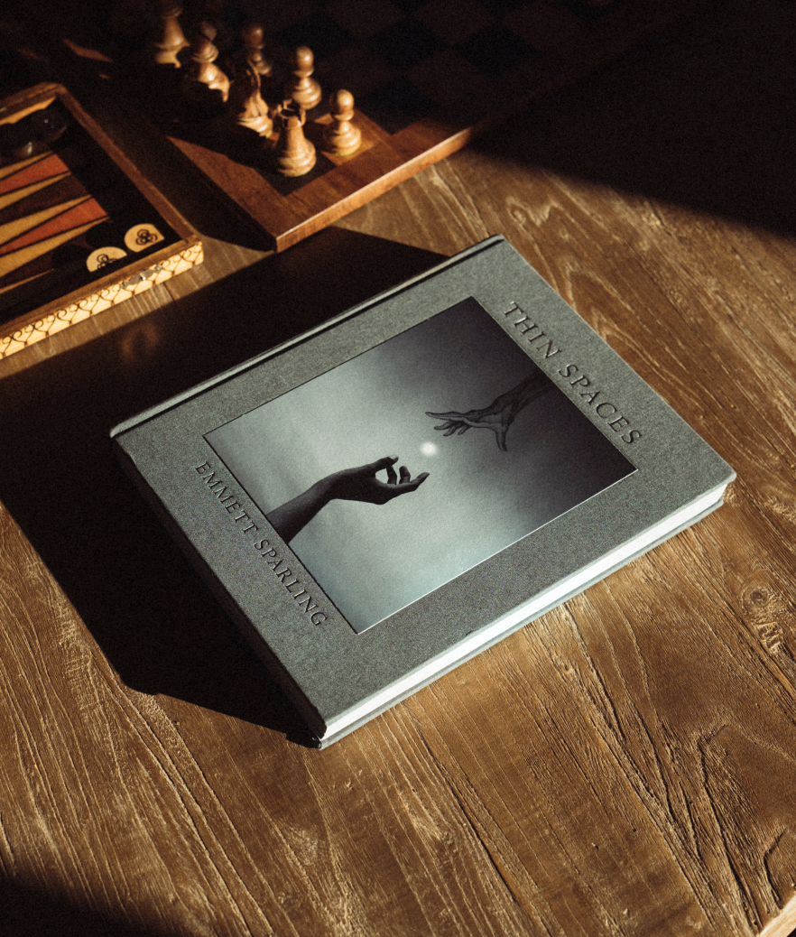 A hardcover book titled "Thin Spaces" by Emmett Sparling lies on a wooden table in dramatic sunlight and shadow, with chess pieces and a backgammon board blurred in the background.