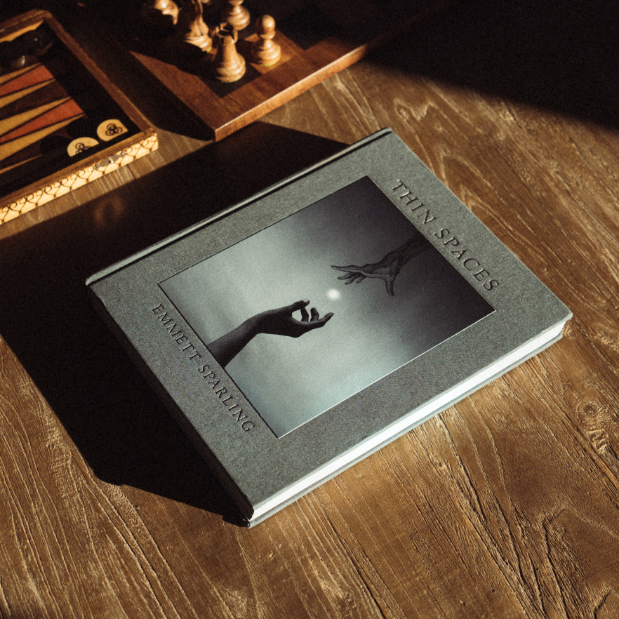 A hardcover book titled "Thin Spaces" by Emmett Sparling lies on a wooden table in dramatic sunlight and shadow, with chess pieces and a backgammon board blurred in the background.