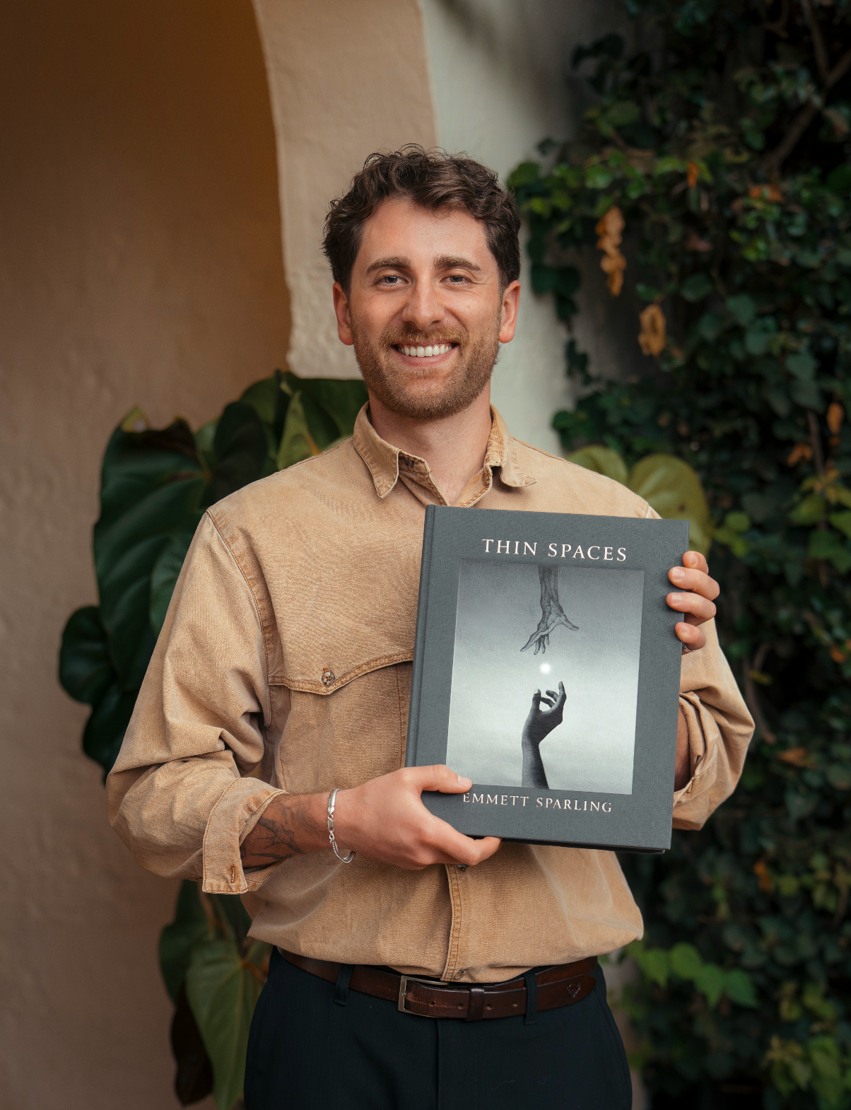 A smiling man in a beige button-up shirt holds up a book titled 'THIN SPACES' by Emmett Sparling. The book cover is dark gray with a black-and-white illustration of a hand holding a glowing light from which a tree grows.
