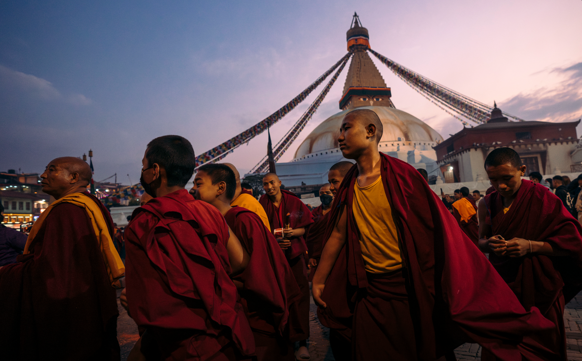 A group of Buddhist monks in maroon and yellow robes walking in front of the large Boudhanwara Stupa in Kathmandu at dusk.