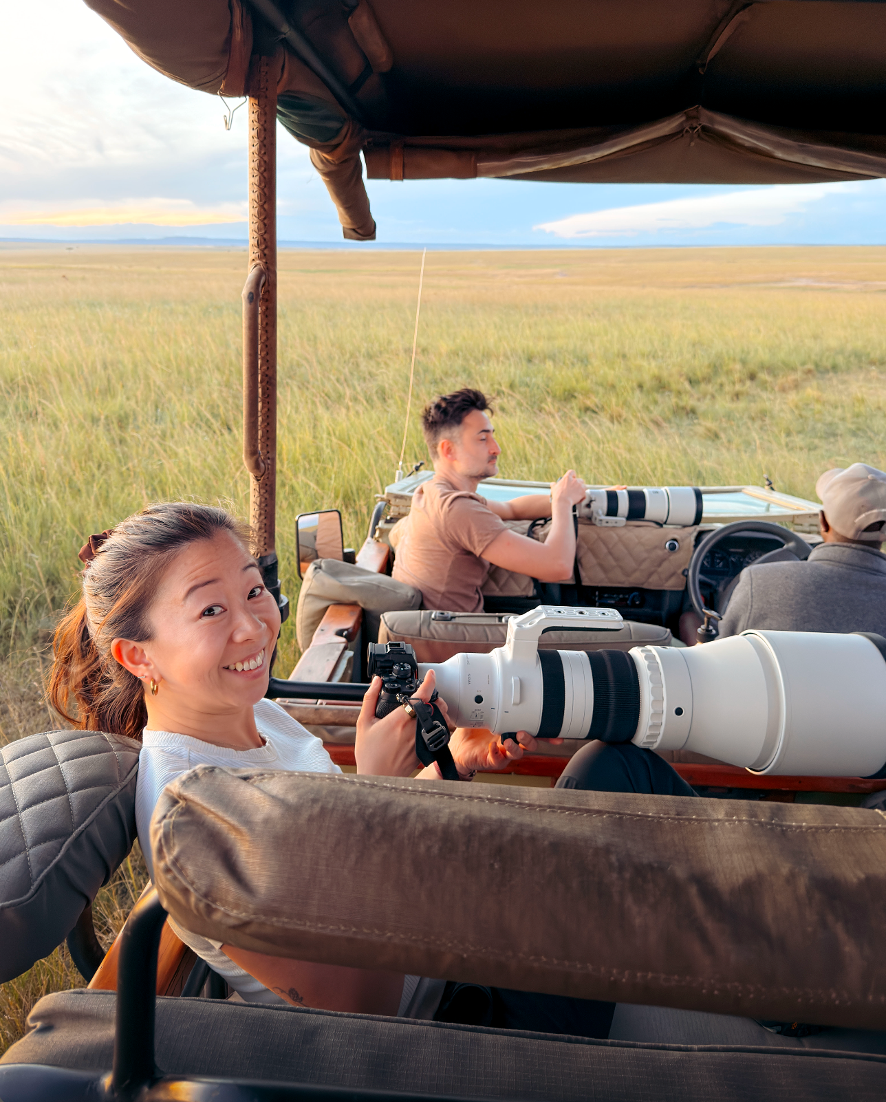 A safari vehicle on a grassy plain with people inside, including a woman in the foreground holding a camera and a man driving. A large white telephoto lens is visible in the front seat.