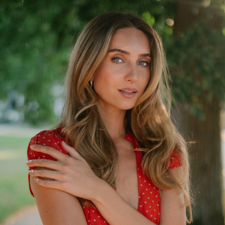A woman with long wavy blonde hair wearing a red sleeveless top with small white polka dots, standing outdoors in front of a blurred green tree background.