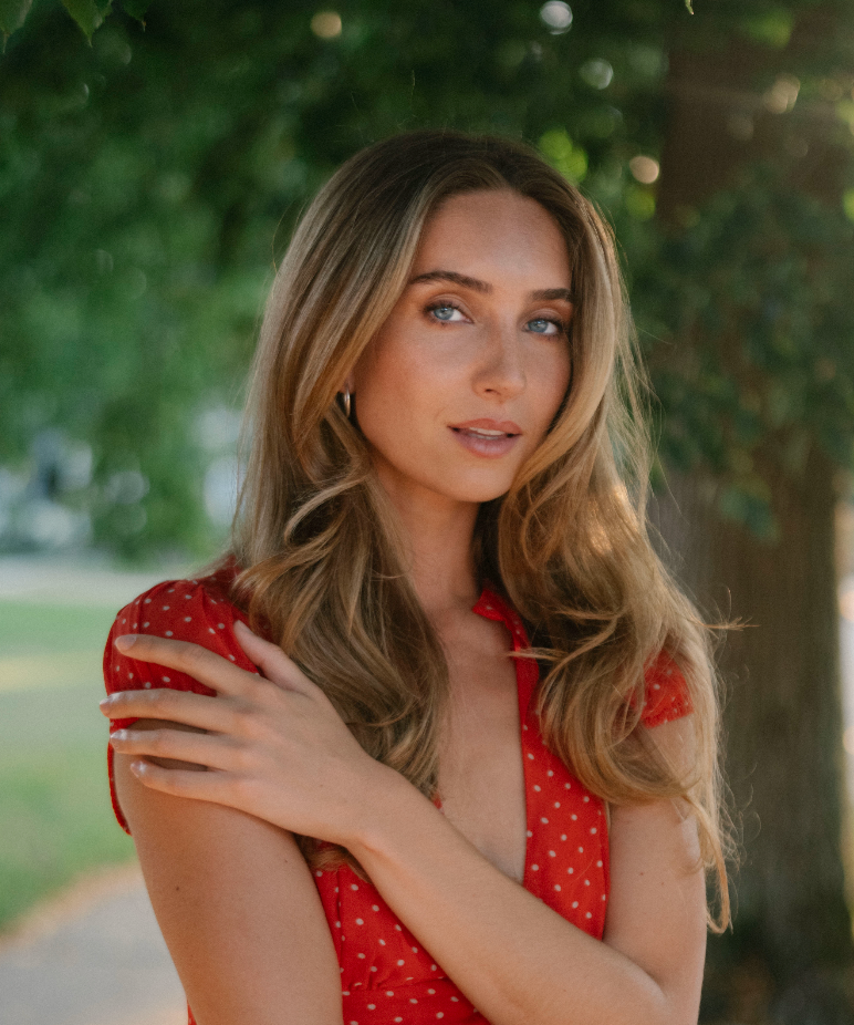 A woman with long wavy blonde hair wearing a red sleeveless top with small white polka dots, standing outdoors in front of a blurred green tree background.